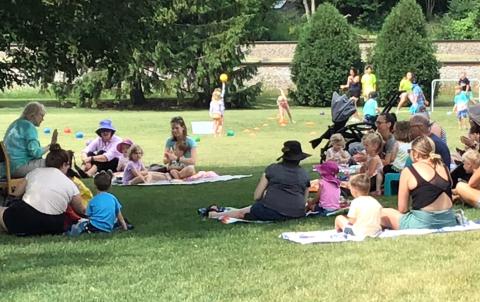 A group of adults and children are gathered outside in the shade of a tree.